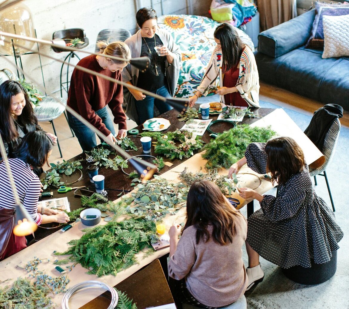 Eine Gruppe junger Frauen ist um einen großen Tisch versammelt, um Kränze zu binden